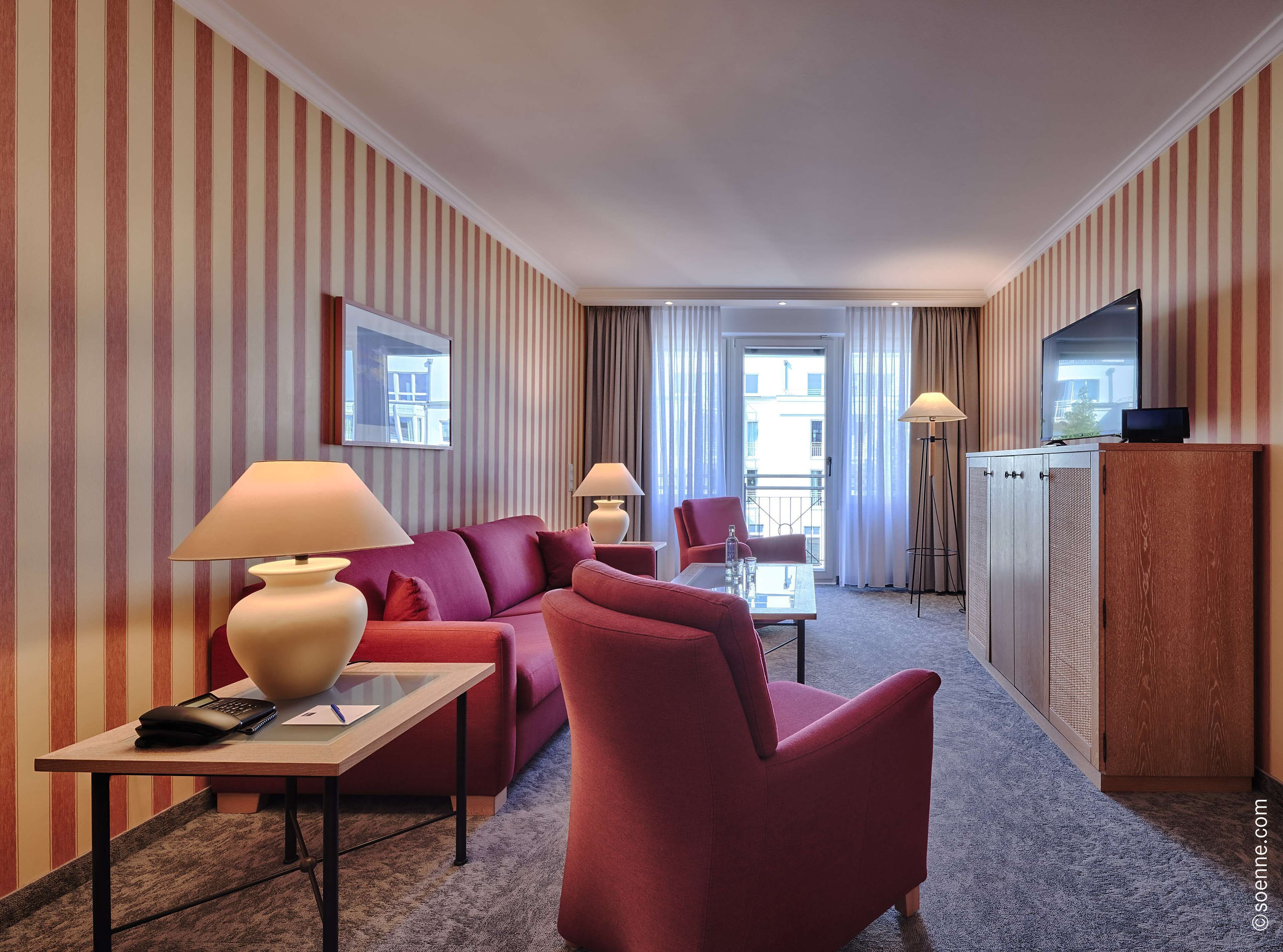 A hotel room with striped wallpaper, a red sofa, a glass table, a desk with a lamp, a chair, a wardrobe, and a TV on top.
