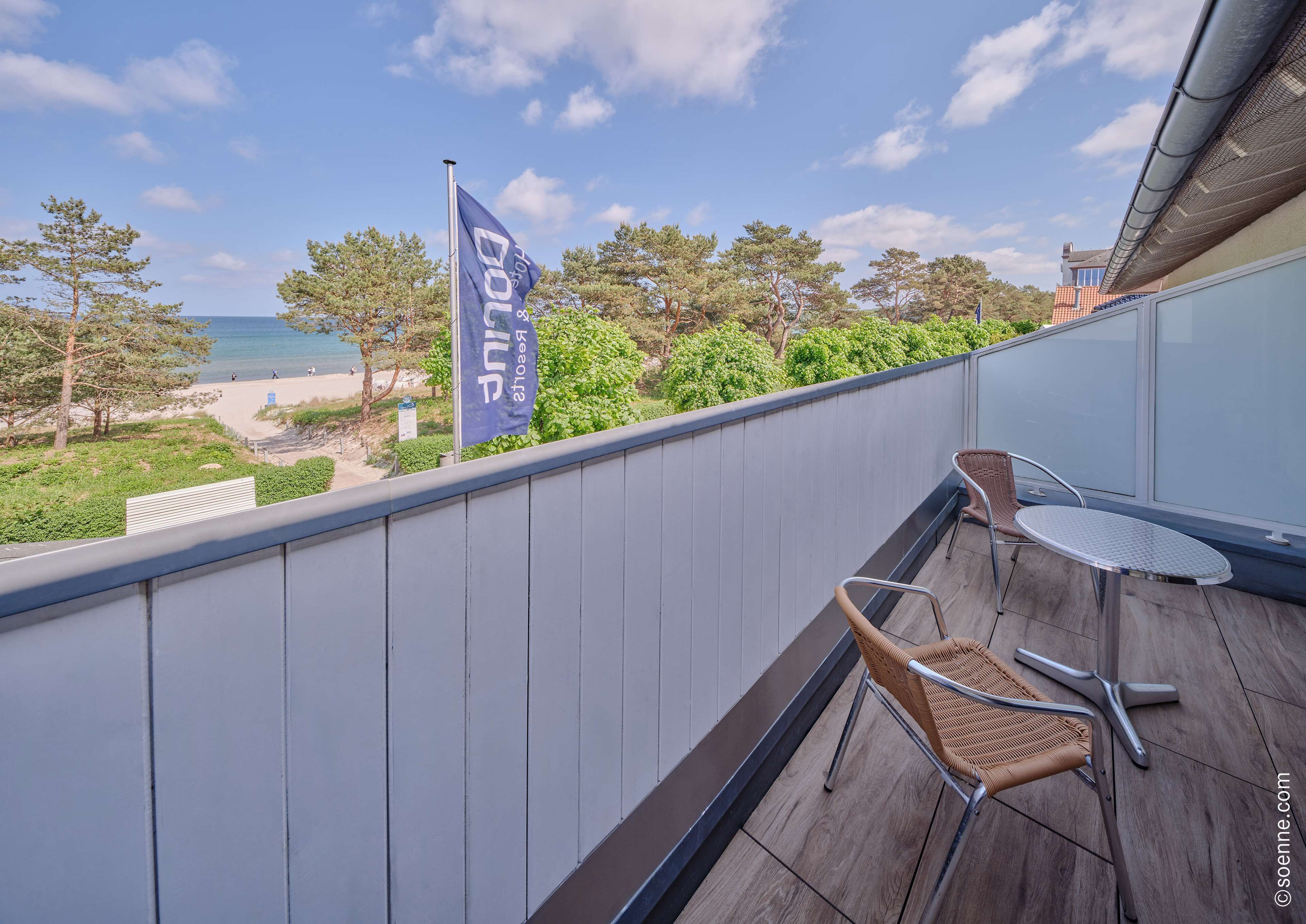 A balcony with two wicker chairs and a small table, overlooking a beach and a flag.