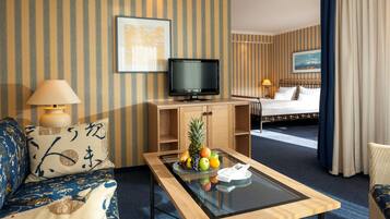 Een hotelkamer met een bed, een televisie op een houten standaard, een glazen bijzettafeltje met fruit, een lamp en een bank met gestreepte kussens.