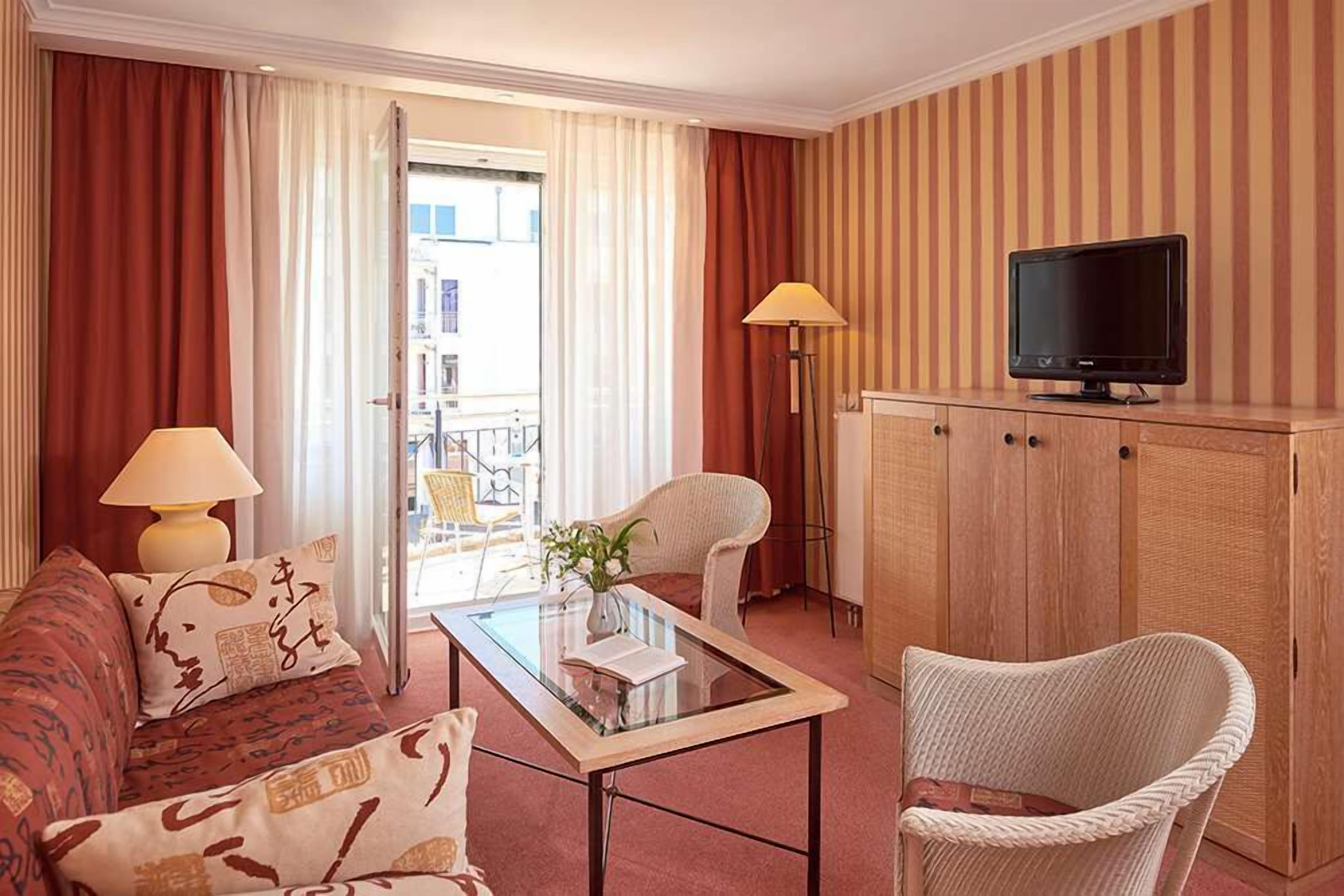 A hotel room with a sofa, a chair, a glass-top coffee table, a television on a wooden cabinet, and a view through a door.