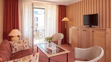 Une chambre d’hôtel avec un canapé, un fauteuil, une table basse en verre, un téléviseur posé sur un meuble en bois et une vue par la porte.