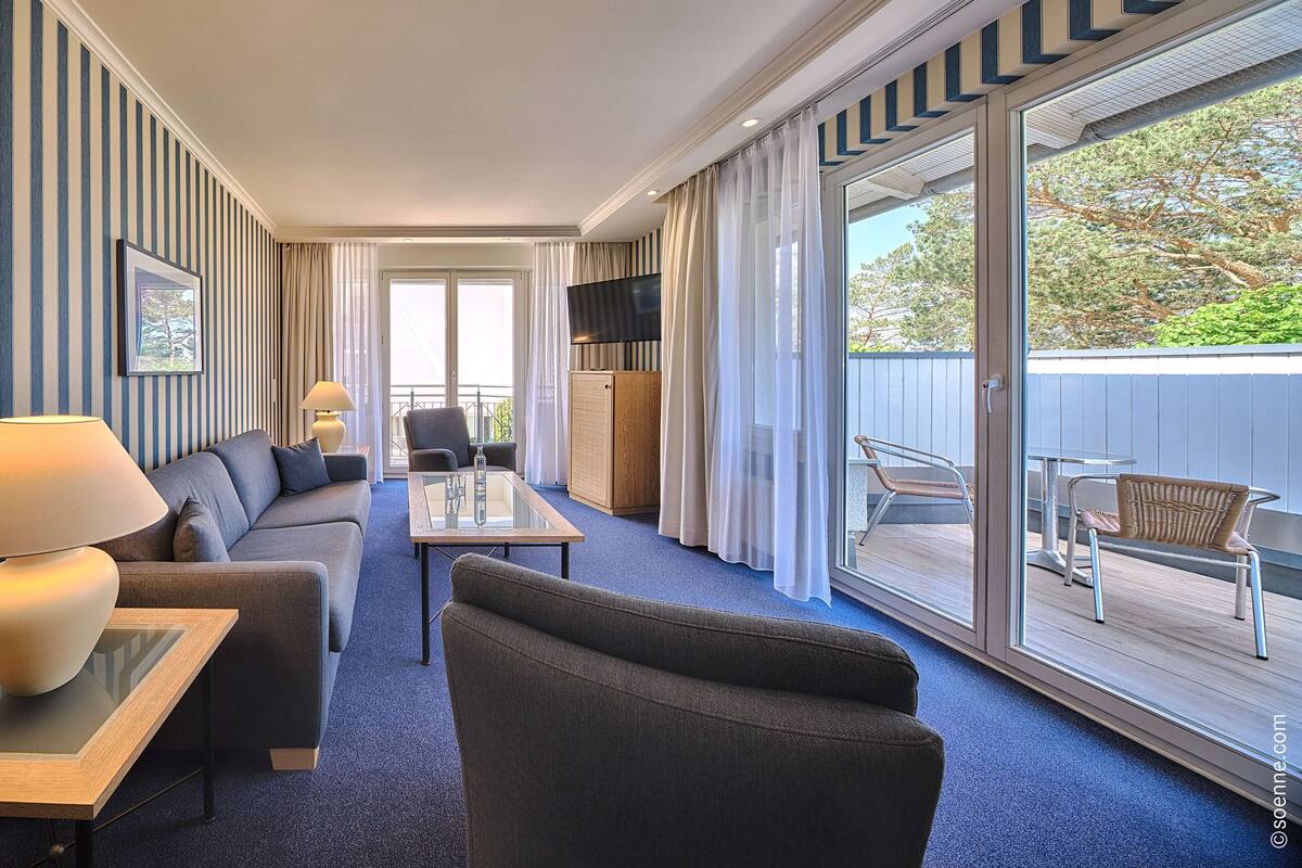 Ein modernes Hotelzimmer mit einer Couch, einem Sessel und einem Glastisch. Ein Fernseher steht auf einer Holzkommode, und es gibt einen Balkon mit Gartenmöbeln.