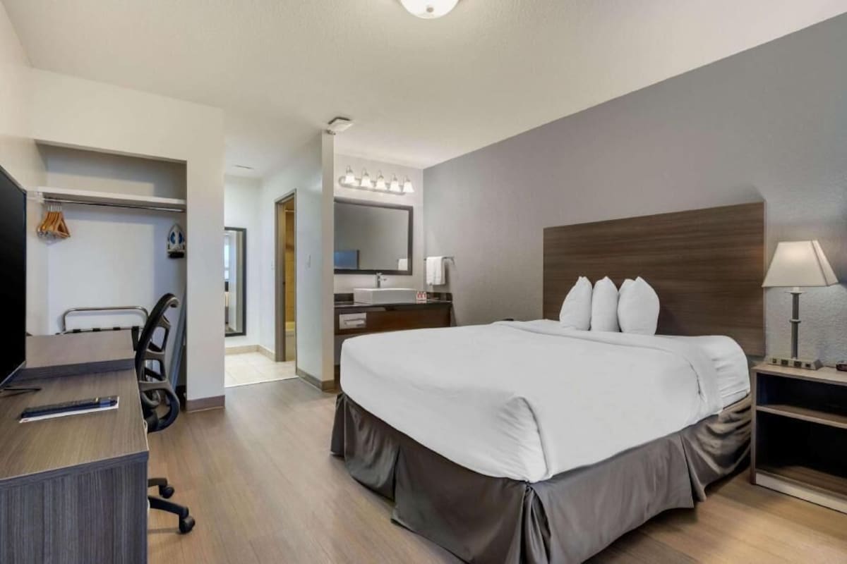 A modern hotel room with a large bed, a desk, a chair, a vanity with a mirror, and a lamp.