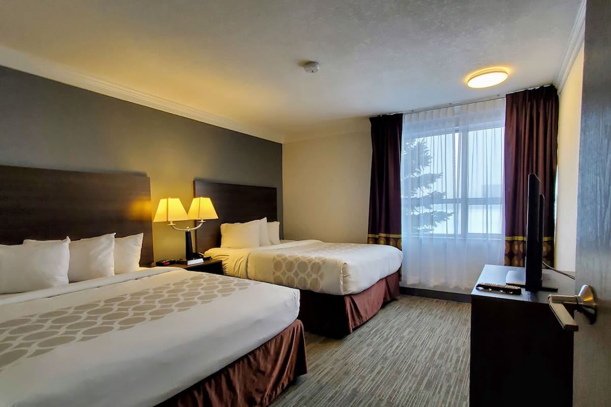 Quarto de hotel com duas camas, uma escrivaninha com computador e controle remoto, uma janela com cortinas e uma luminária no teto.