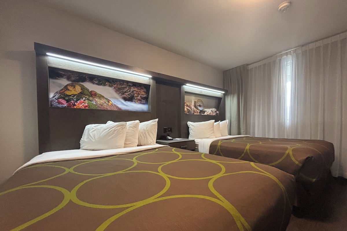 Quarto de hotel com duas camas, cada uma com cabeceira e uma janela ampla. Há uma televisão de tela plana fixada na parede acima das camas.