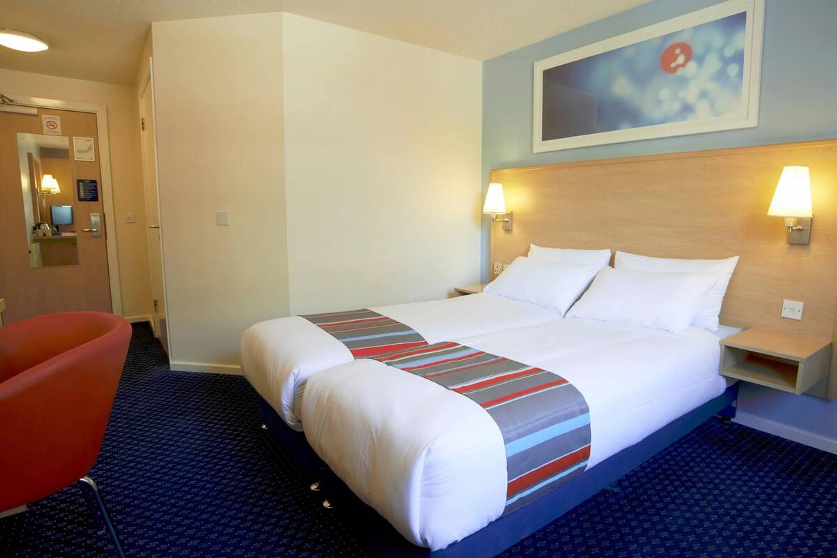 A hotel room with a bed, a red chair, a nightstand, and a wall-mounted lamp.
