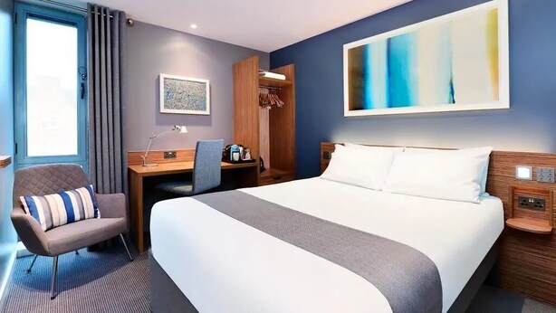 A hotel room with a large bed, a desk, a chair, and a wardrobe. - Travelodge London Docklands (London)