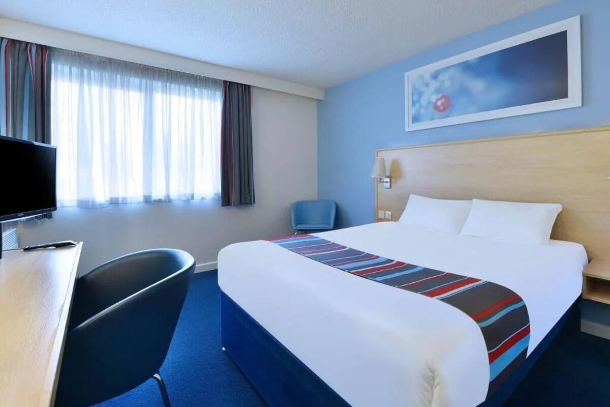 A hotel room with a bed, a desk, a chair, a TV, and a painting on the wall.