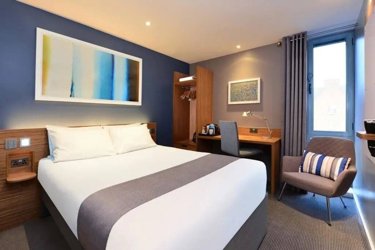 A hotel room with a large bed, a desk, a chair, and a window with curtains.