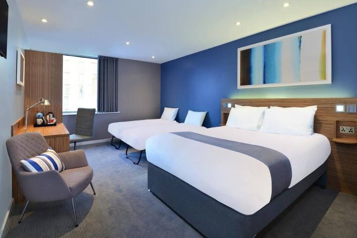 A modern hotel room with two beds, a seating area, and a large window.