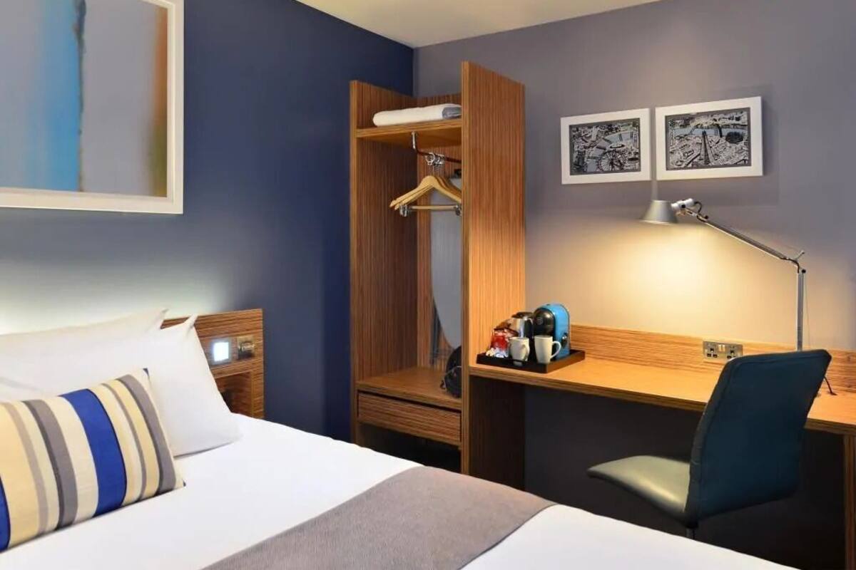 A hotel room with a bed, a desk, a chair, a wardrobe, a bedside table, and two framed pictures on the wall.