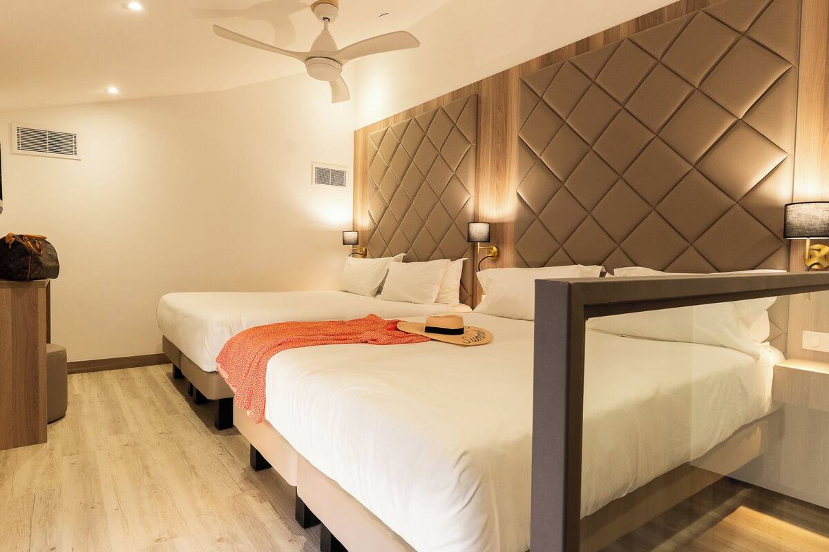 Una habitación de hotel con cama, lámparas de noche, ventilador de techo y cabecero de madera.