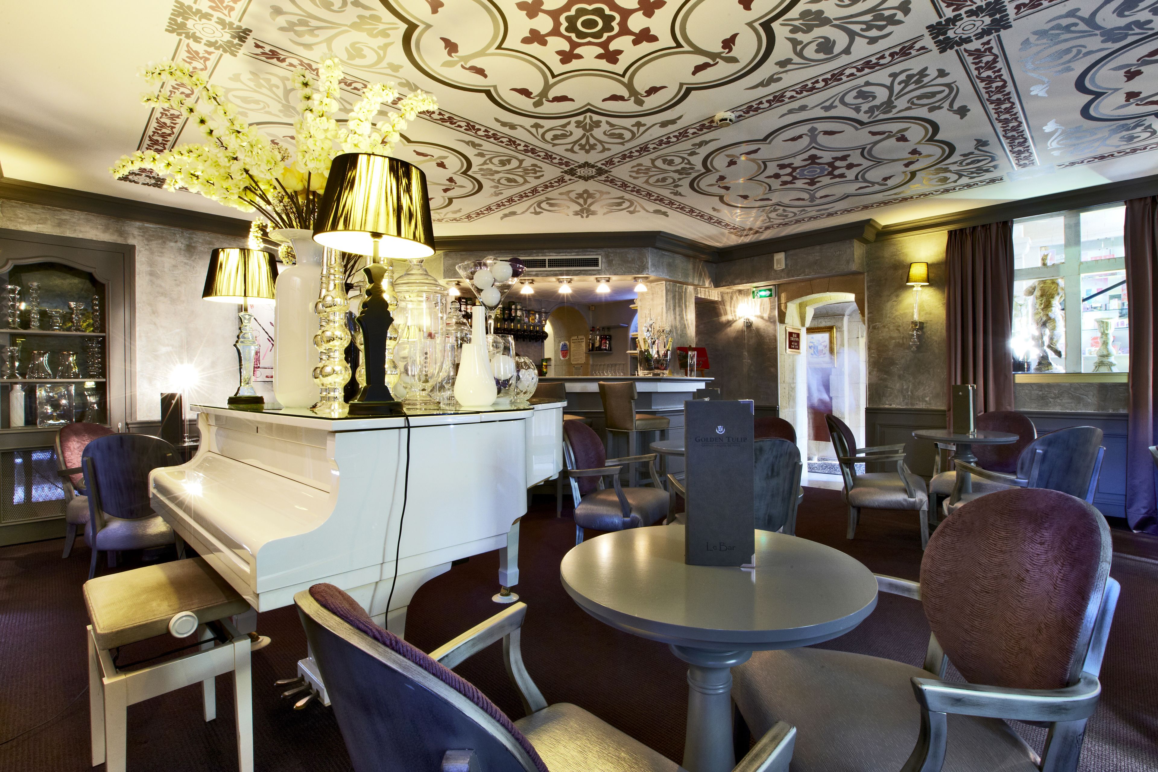 A room with a piano, round tables, and chairs, featuring a decorative ceiling and a bar area with bottles and glasses.