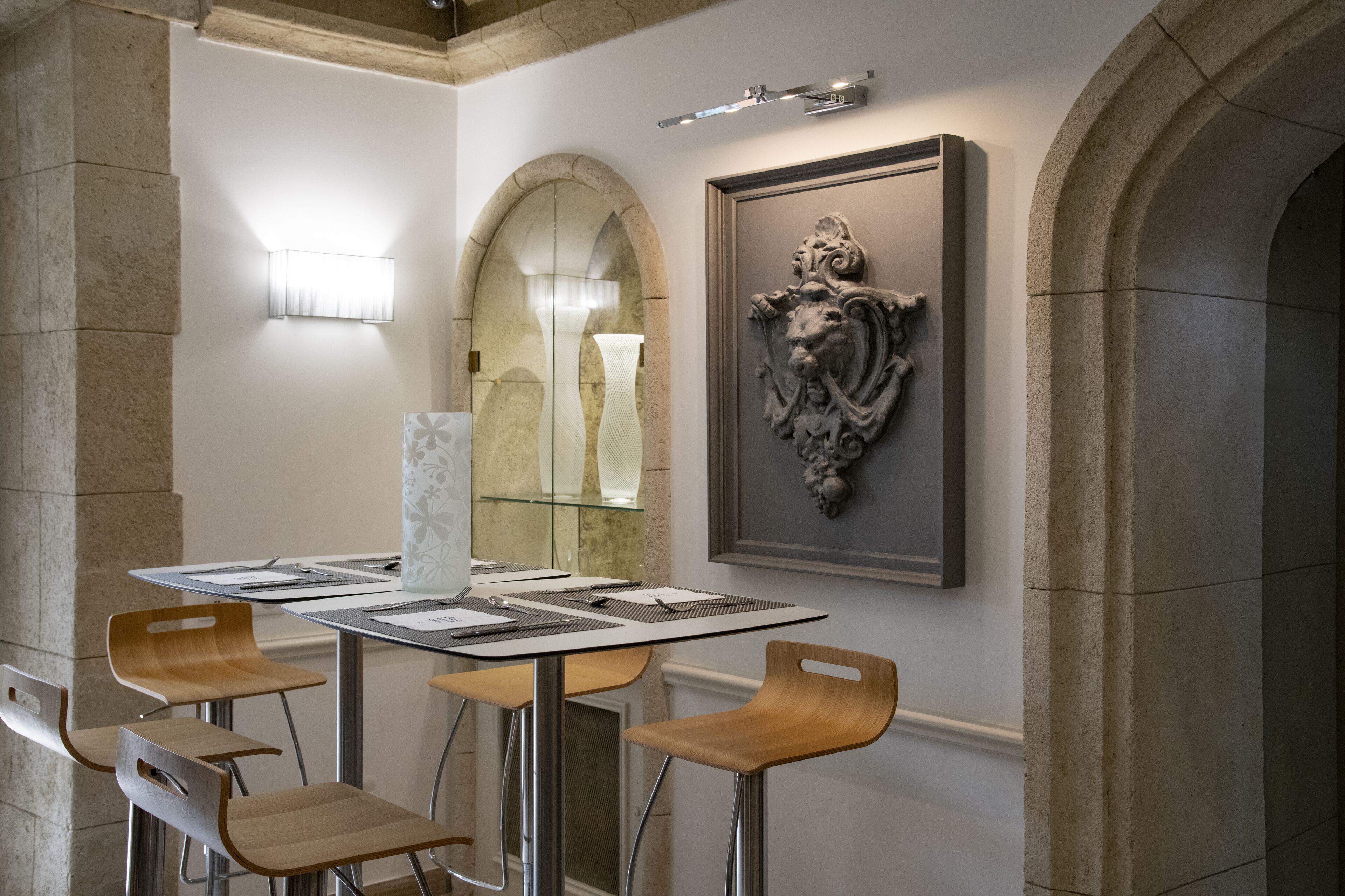 A modern dining area with a table set for four, featuring a vase centerpiece, a framed artwork above the table, and a stone column to the left.