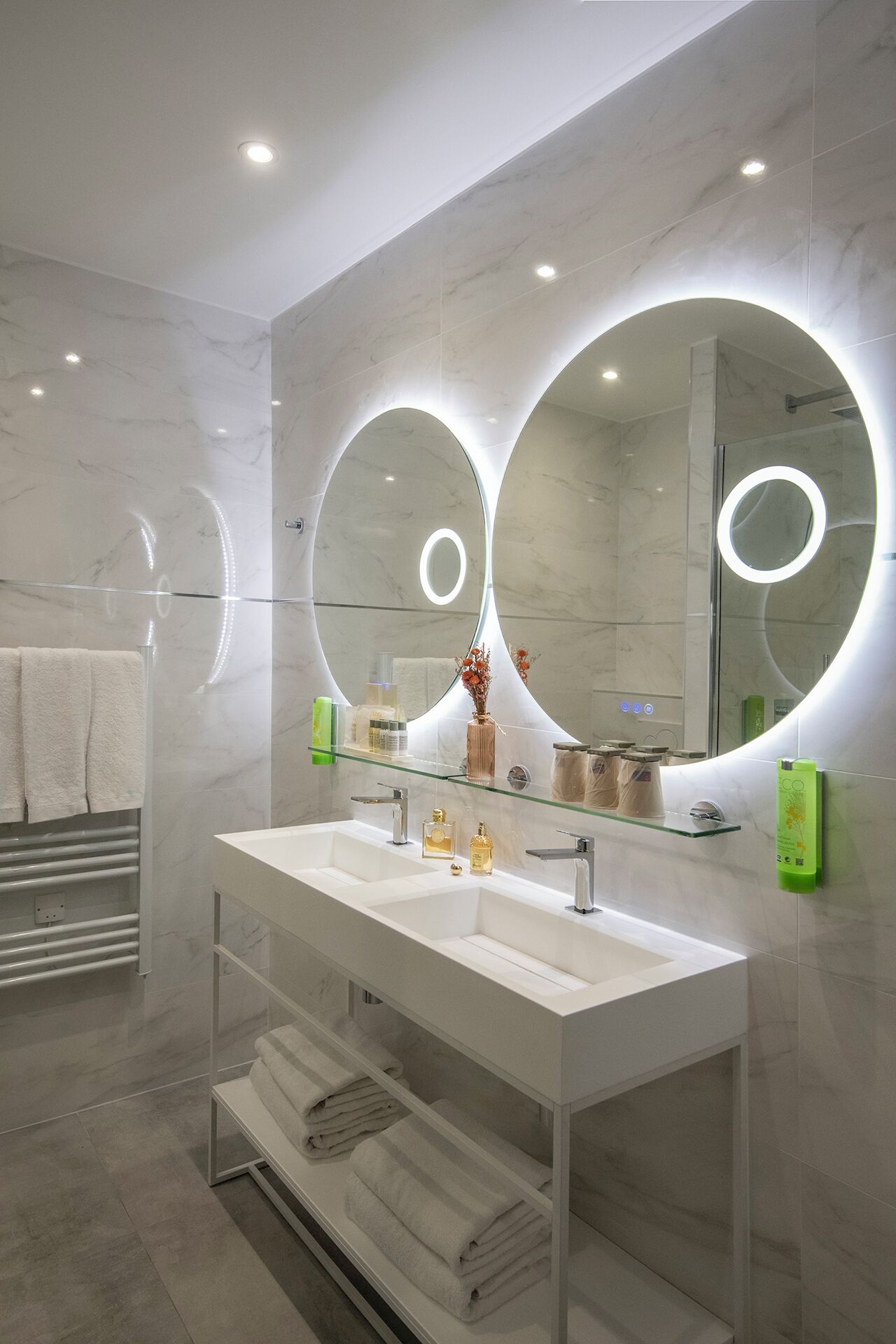 A modern bathroom with a double sink, large mirrors, and a glass shelf with toiletries.