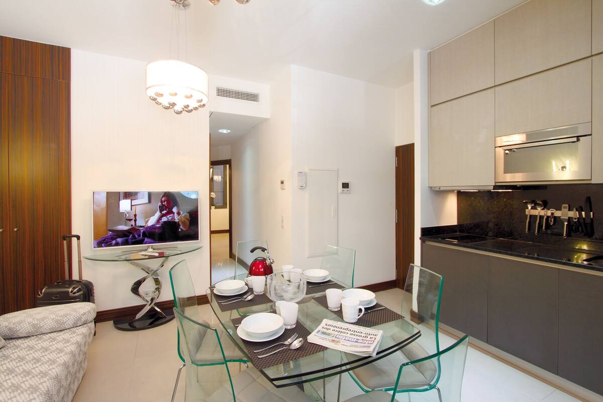 Una habitación de hotel moderna con una mesa de comedor de vidrio, un sofá, un televisor y una zona de cocina.