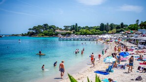 A beach with clear turquoise water, people swimming and sunbathing, and a pier with boats. - Golden Tulip Cannes hotel de Paris (Cannes)