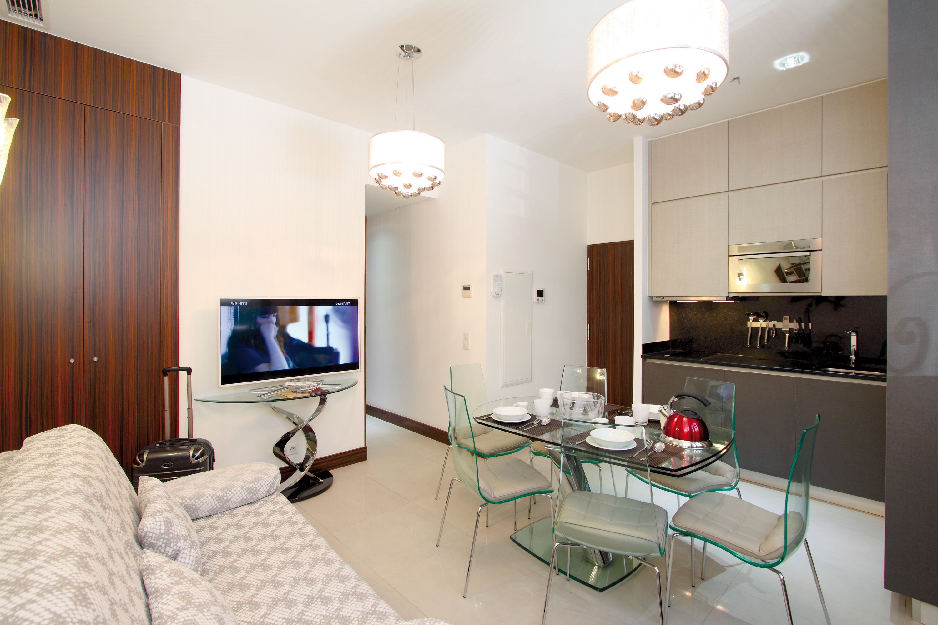 A modern living room with a sofa, a glass dining table with chairs, a television, and a kitchen area with cabinets and a stove.