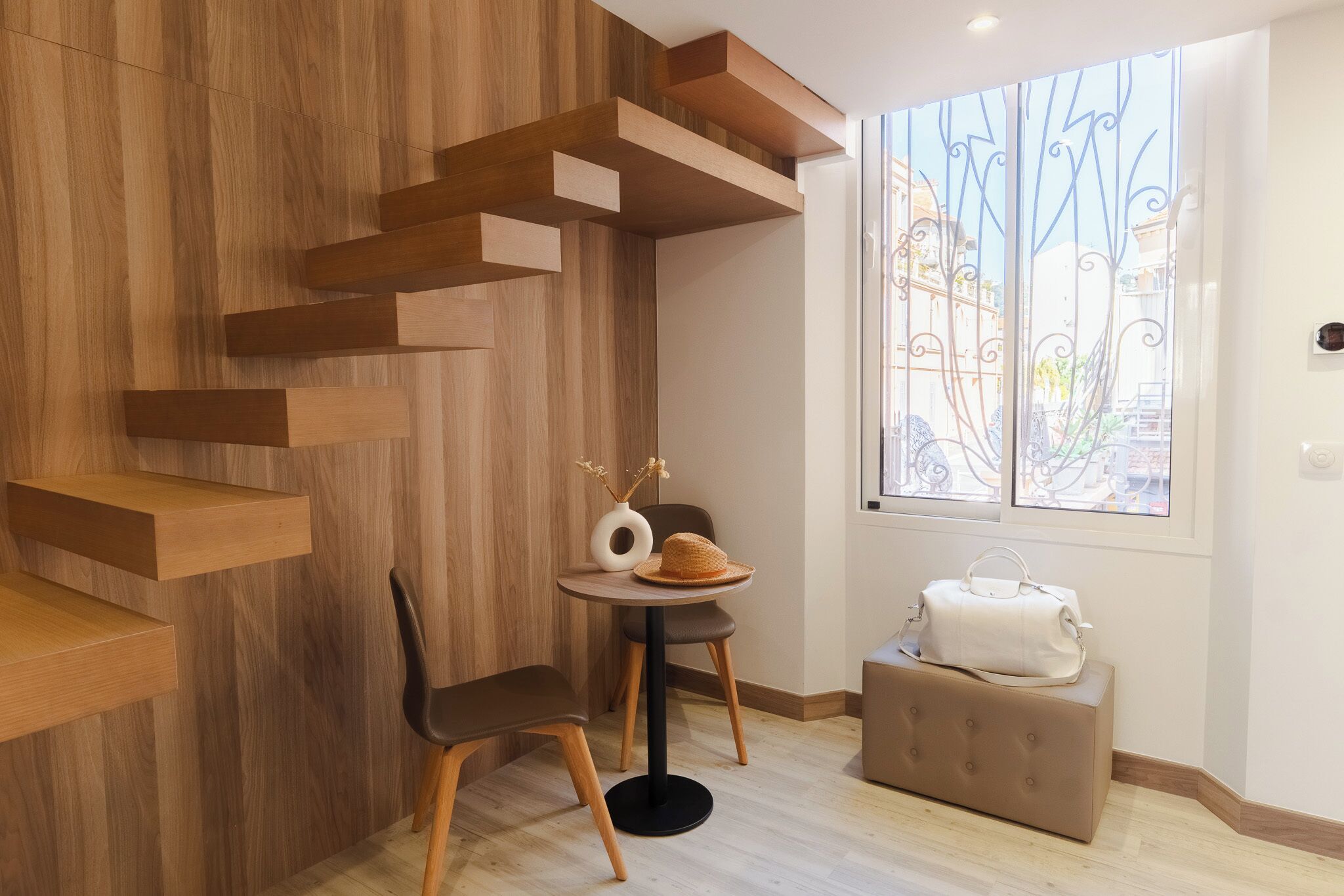 A modern room with a wooden wall, a window with decorative iron bars, a round table with a vase and a hat, a chair, and a suitcase.