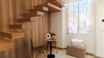 A modern room with a wooden wall, a window with decorative iron bars, a round table with a vase and a hat, a chair, and a suitcase.