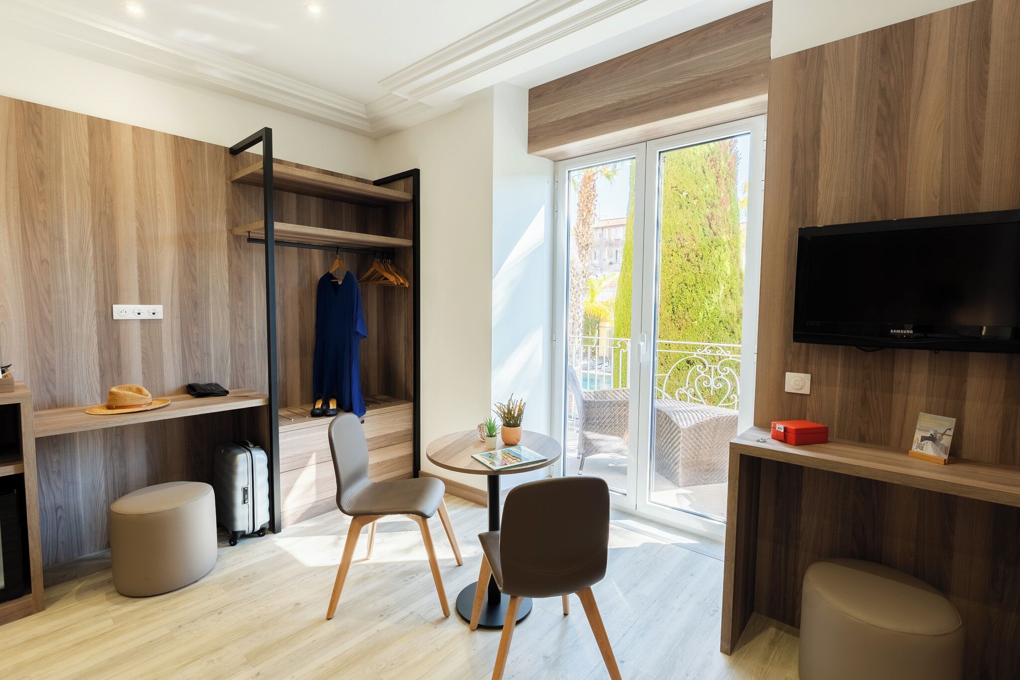 Een moderne hotelkamer met een houten vloer, een balkon met tuinmeubilair, een aan de muur bevestigde televisie en een bureau met een stoel.