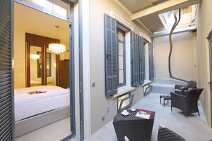 Een hotelkamer met een bed, een tafeltje en een stoel. Er zijn ramen met luiken en een kroonluchter.