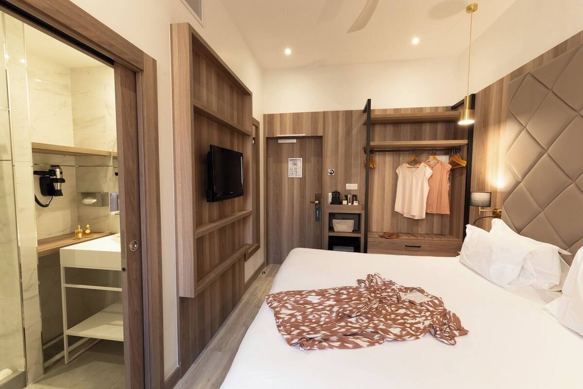 Una habitación de hotel con una cama, televisor, armario de madera, espejo, tocador con lavabo y un perchero.