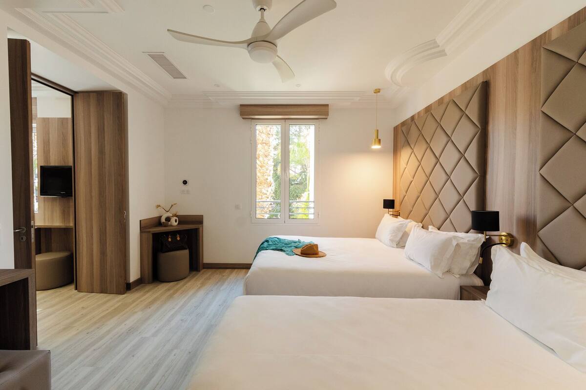 Habitación de hotel con dos camas, ventilador de techo, televisor, ventana con persianas y un panel de pared de madera.