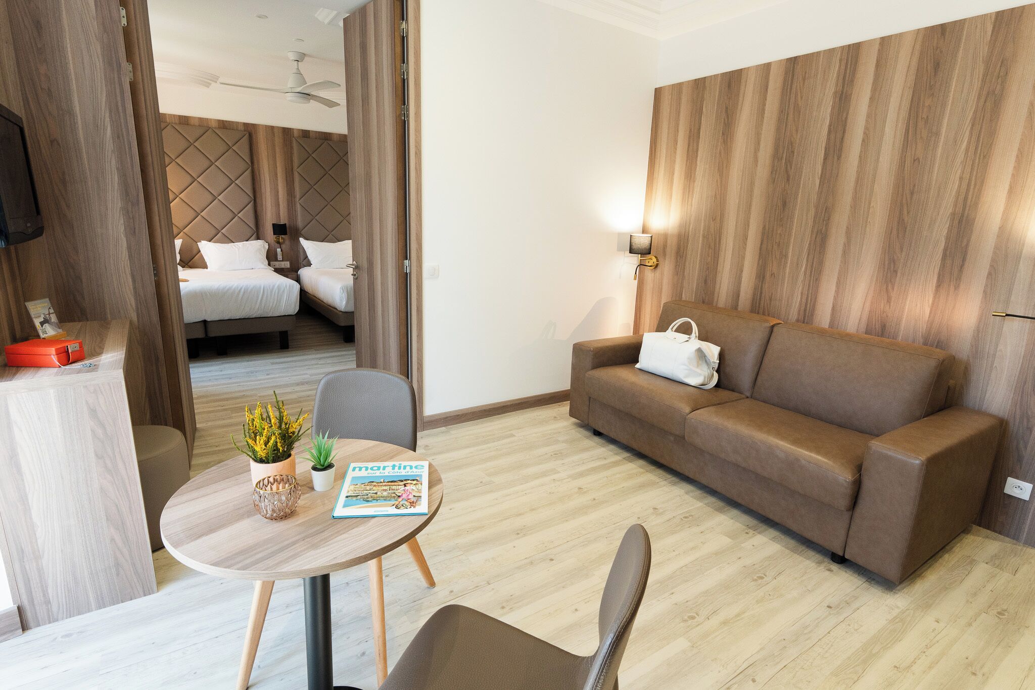 A hotel room with a bed, a sofa, a dining table with chairs, a small plant, and a magazine.