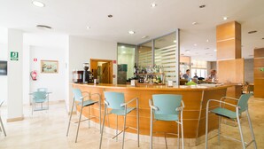 Un área de bar moderna con mostrador de madera y sillas de metal. Hay varias botellas y una máquina de café en el mostrador.