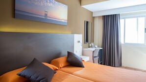 Habitación de hotel con cama, escritorio, silla, un cuadro de un atardecer y una ventana con cortinas.
