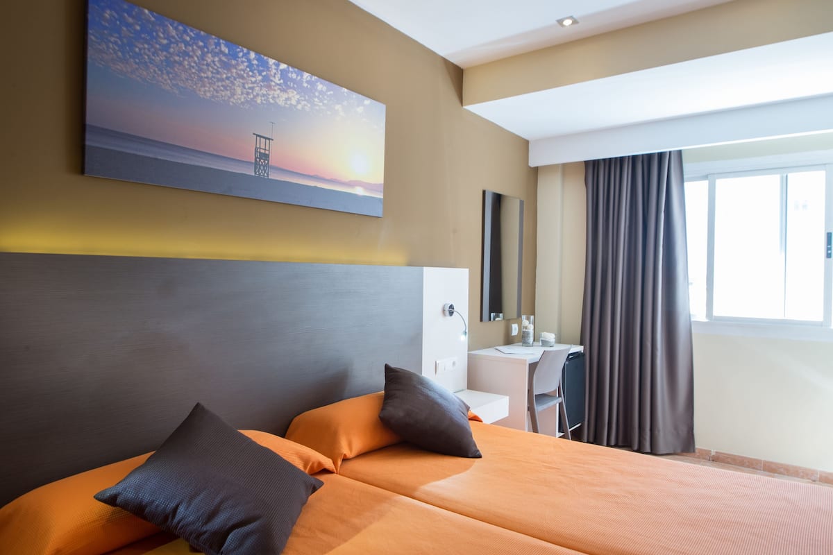 Habitación de hotel con cama, escritorio, silla, un cuadro de un atardecer y una ventana con cortinas.
