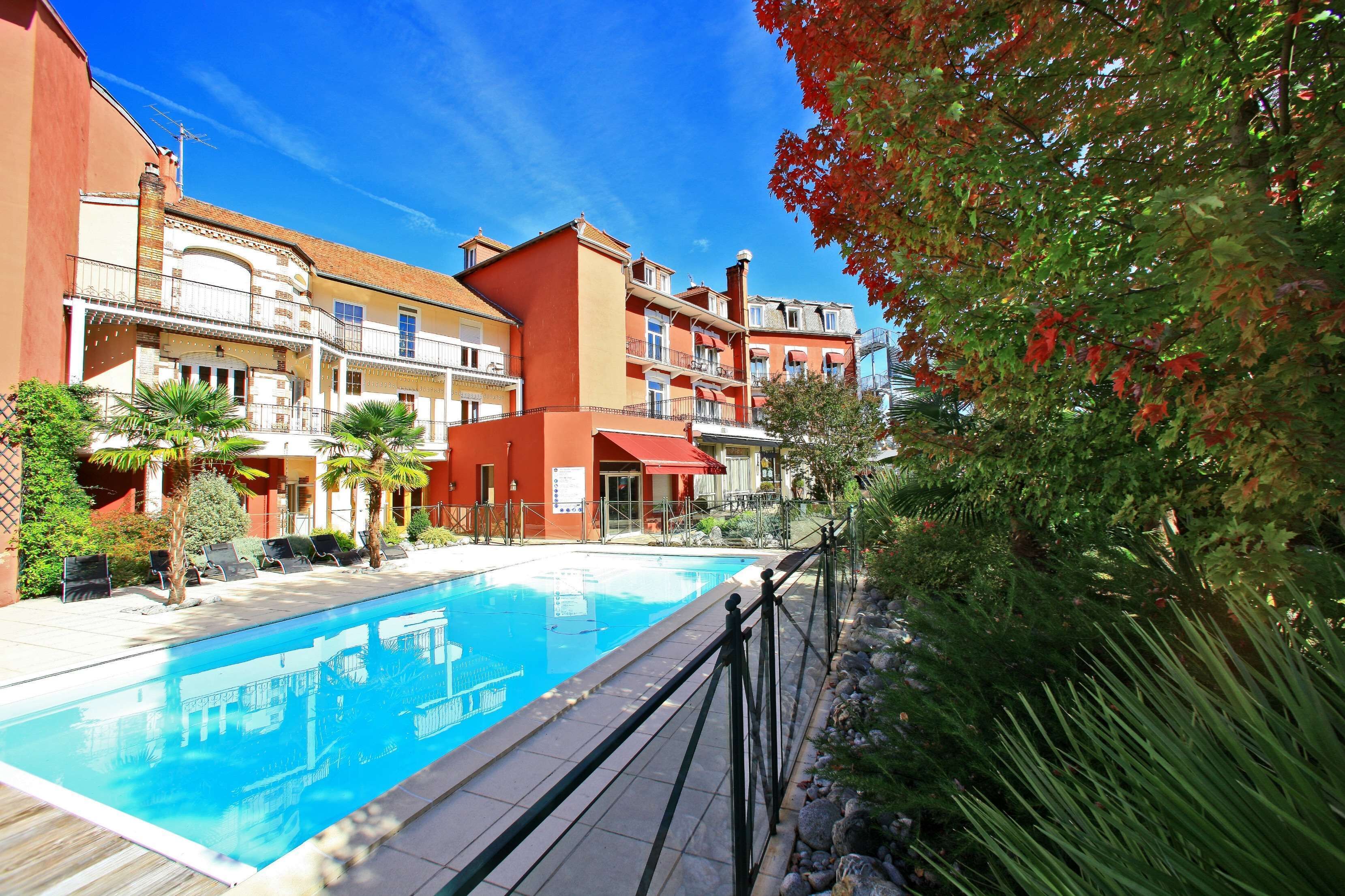 A swimming pool surrounded by multi-story buildings with balconies, a clear blue sky, and some greenery.