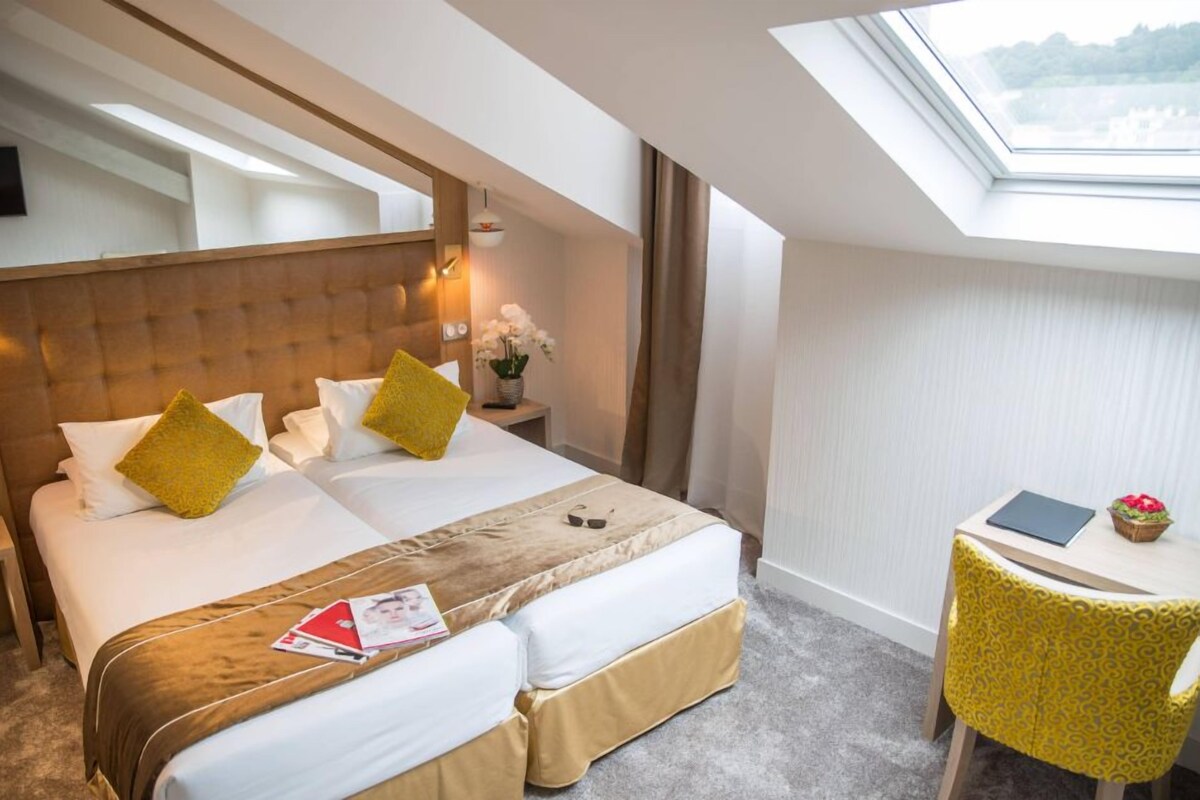 Habitación de hotel con una cama grande, dos almohadas, una mesita de noche con una lámpara, una pequeña mesa con una computadora portátil y flores, y una ventana con vistas.