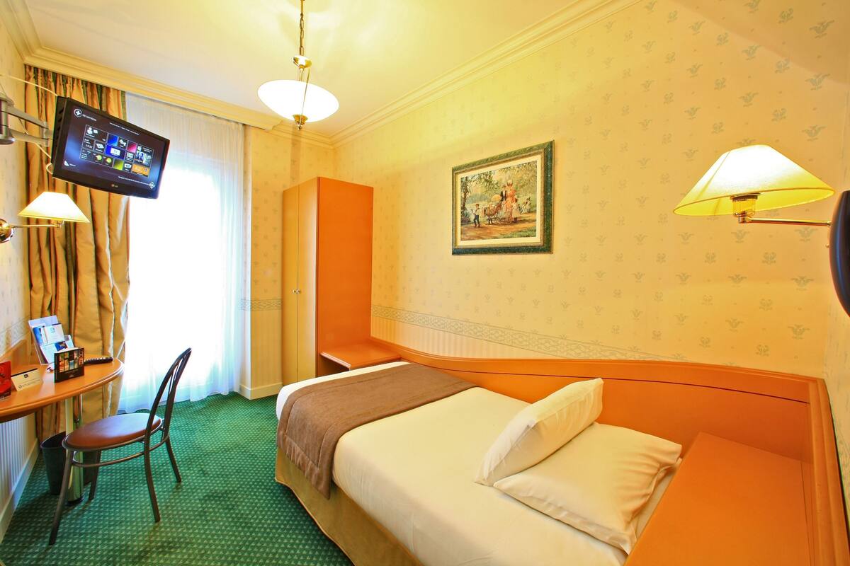 Une chambre d’hôtel avec un lit, un bureau avec un ordinateur, une chaise, un téléviseur fixé au mur et un tableau accroché au mur.