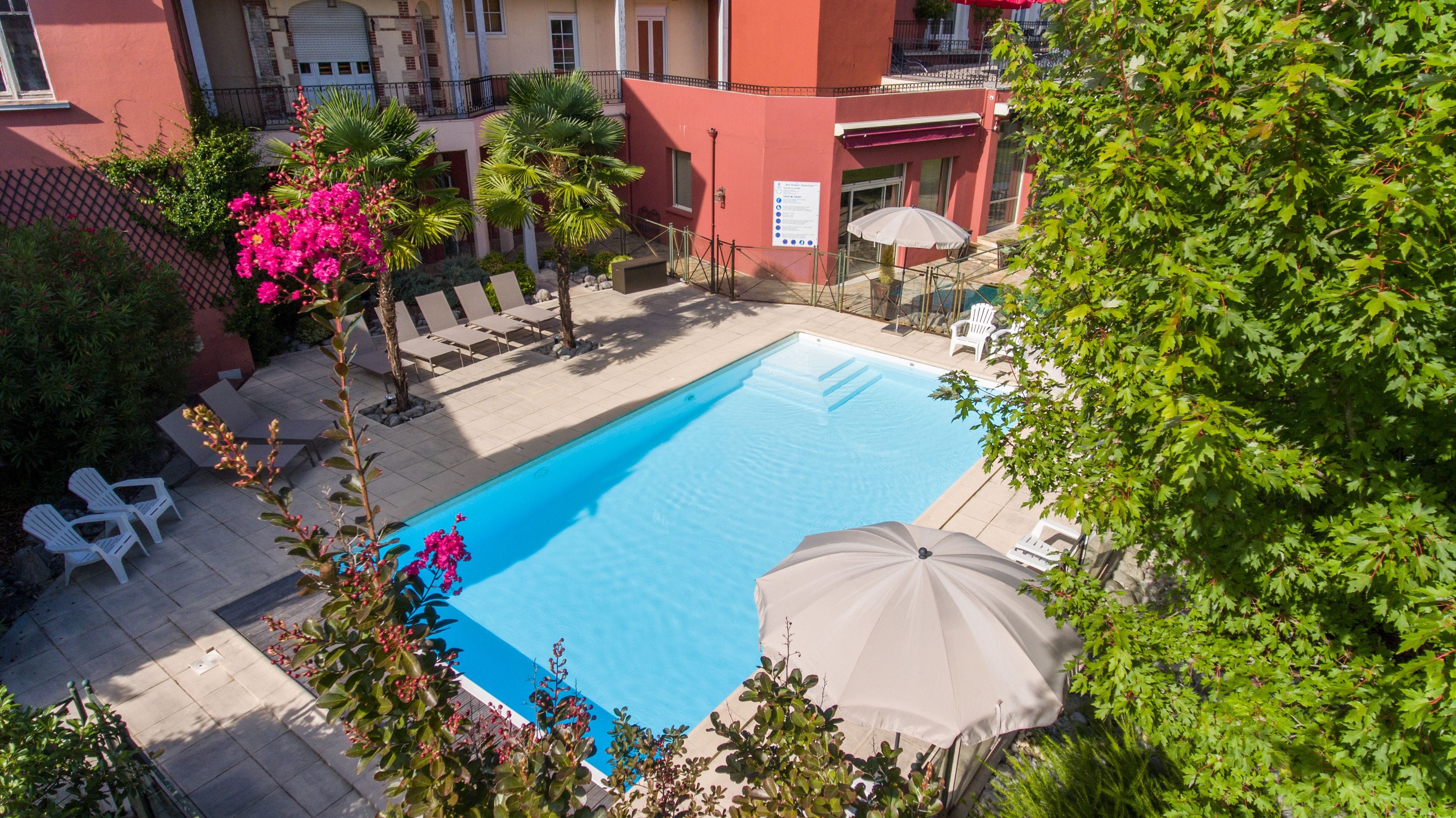 A swimming pool with lounge chairs and an umbrella, surrounded by greenery and a building with a red facade.
