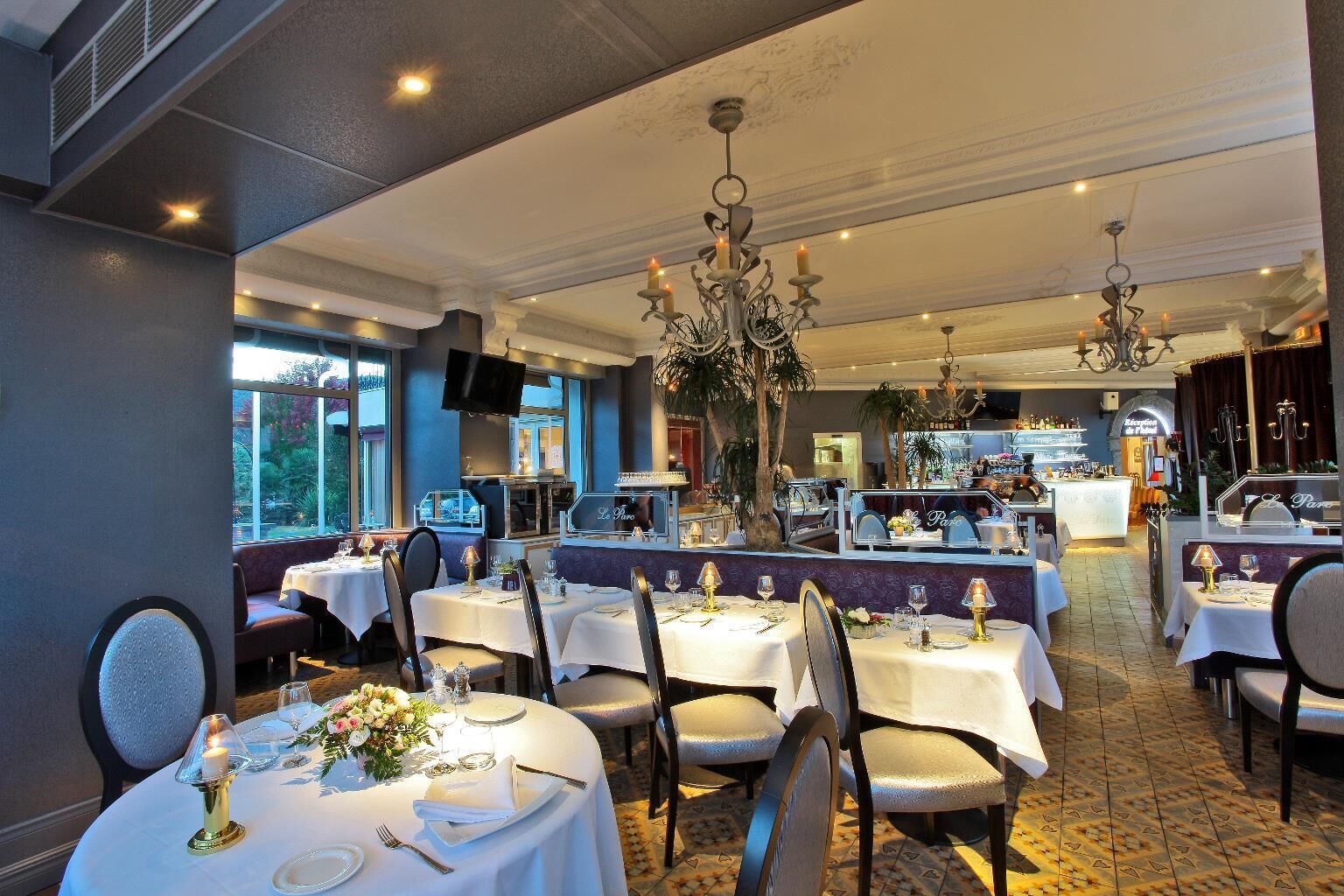 A restaurant with round tables set for dining, a bar area in the background, and a large chandelier.