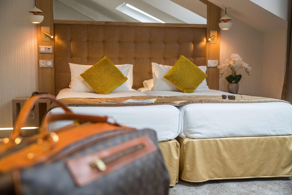 Une chambre d’hôtel avec un grand lit, deux oreillers, une table de chevet, une lampe, un vase avec des fleurs et une valise posée sur le sol.