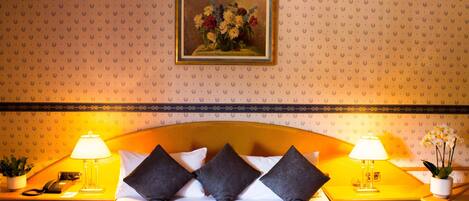 A hotel room with a large bed, two bedside tables with lamps, a telephone, a potted plant, and a framed floral painting on the wall.