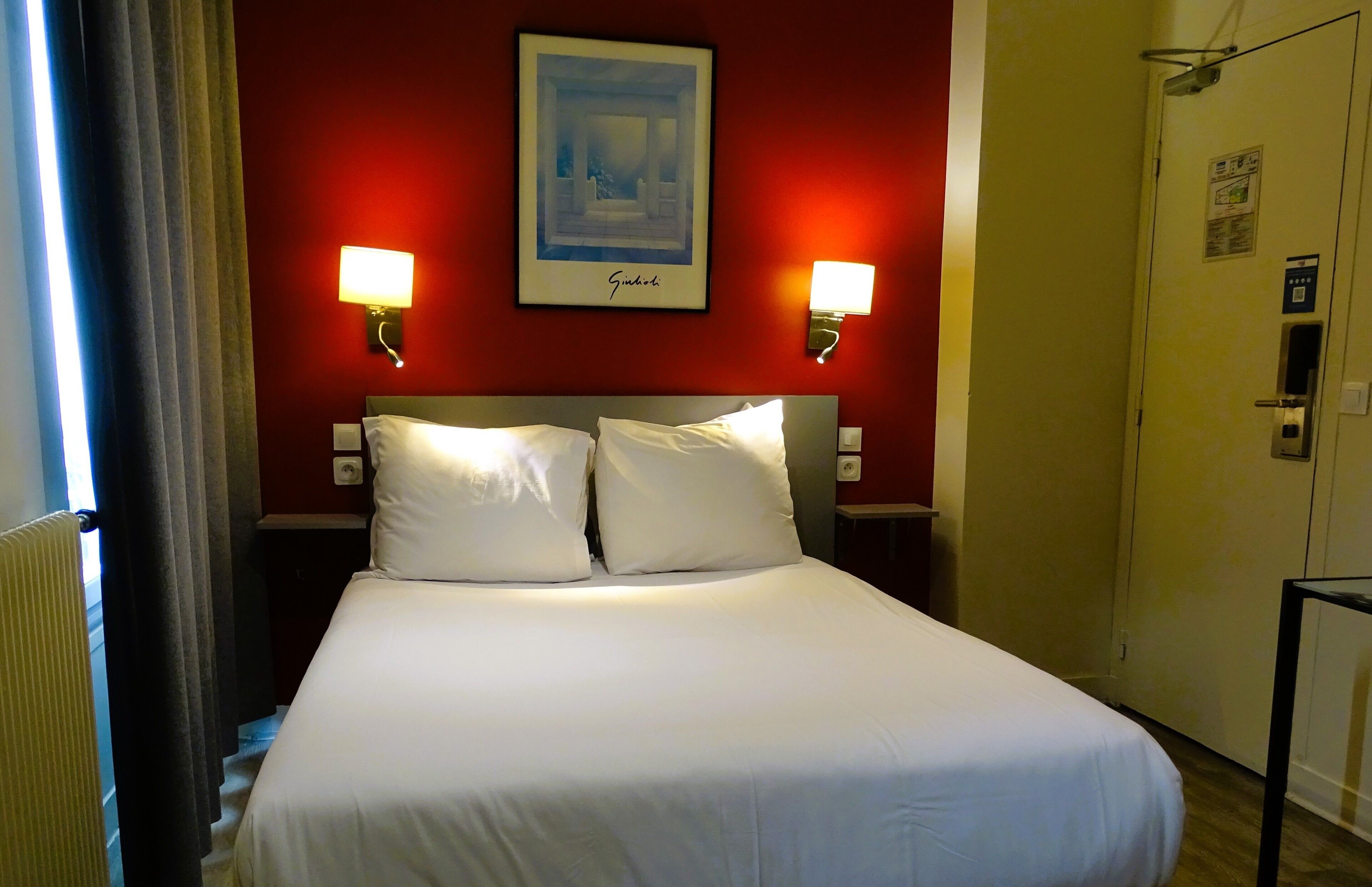 A hotel room with a large bed, two bedside lamps, a framed picture on the wall, and a door with an elevator button.