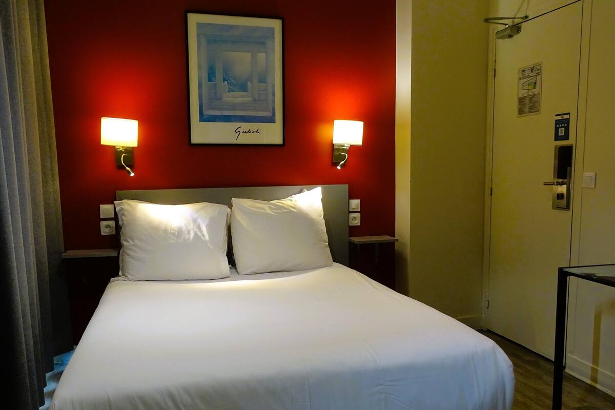A hotel room with a bed, two bedside lamps, a framed picture on the wall, and a door with a lock and a light switch.