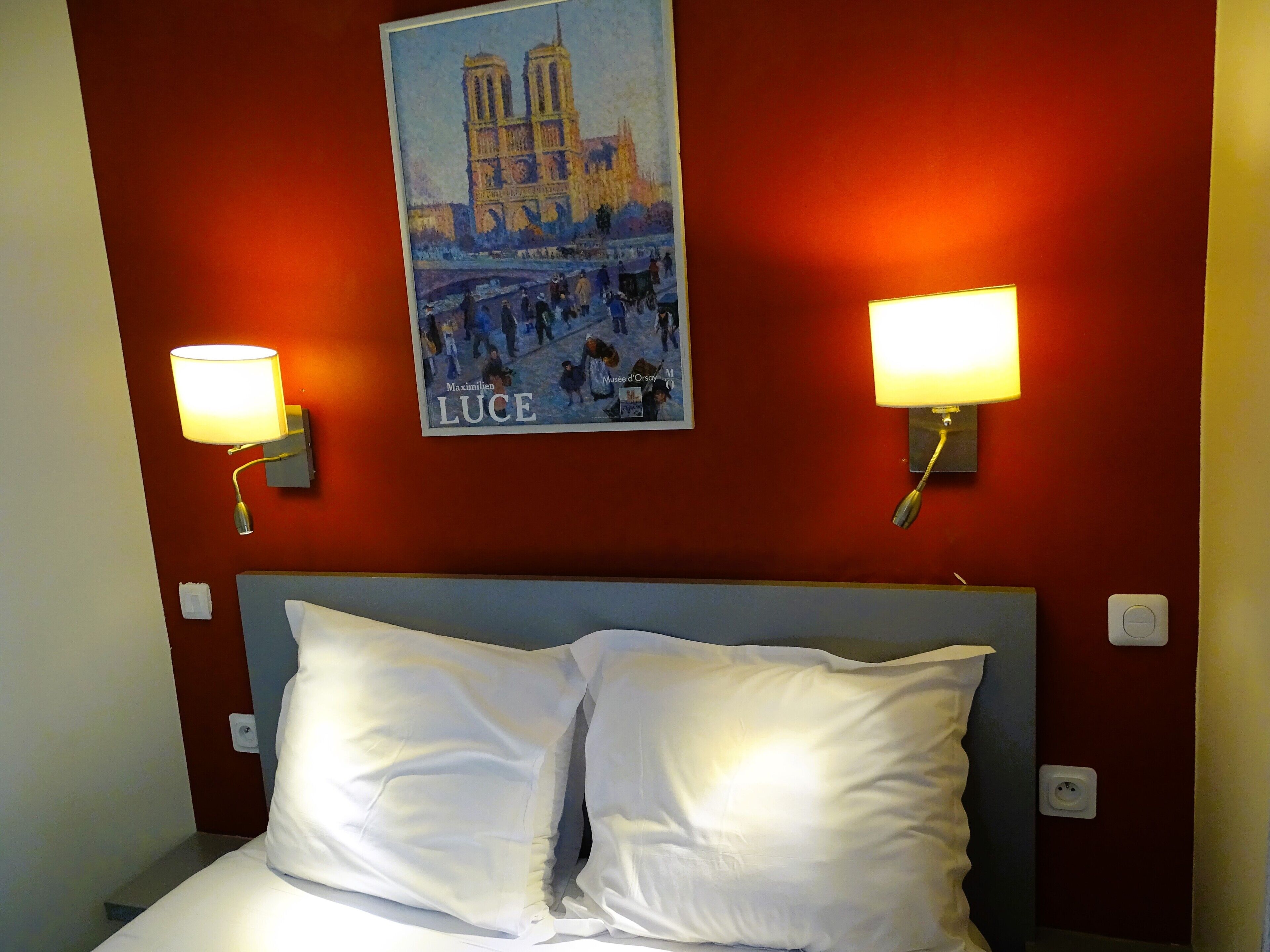 A hotel room with a bed, two wall-mounted lamps, and a poster of Notre-Dame Cathedral.