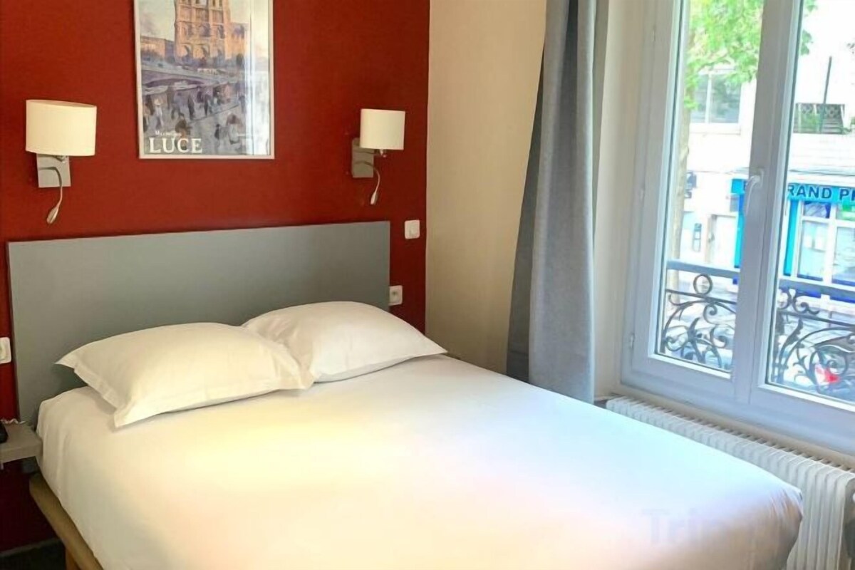 A hotel room with a bed, two wall-mounted lamps, a poster, and a window with a view of the street.