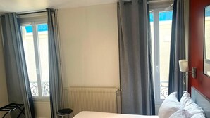 Una camera d'albergo con un letto, un termosifone, una finestra con tende e un piccolo sgabello, con vista all'esterno.