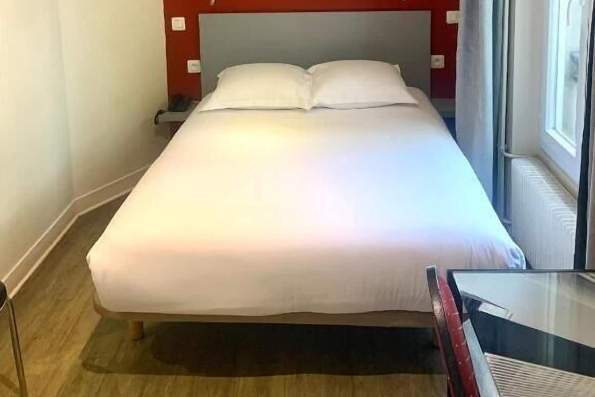 A hotel room with a single bed, a red accent wall, a poster of a cathedral, two bedside lamps, and a window with curtains.
