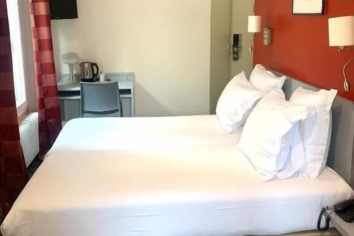 A double bed with white bedding, a desk with a chair, a kettle, and a flat-screen TV on the wall.