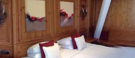 A wooden-paneled room with a bed, two paintings, and wall sconces.