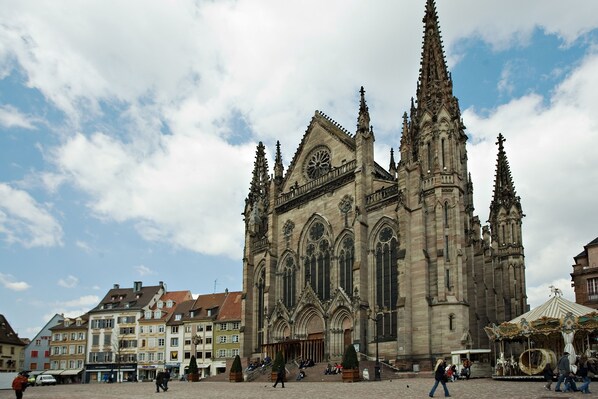 A historic cathedral with intricate Gothic architecture, a prominent clock tower, and a bustling square with pedestrians and a market stall. - Hotel Bristol (Mulhouse)