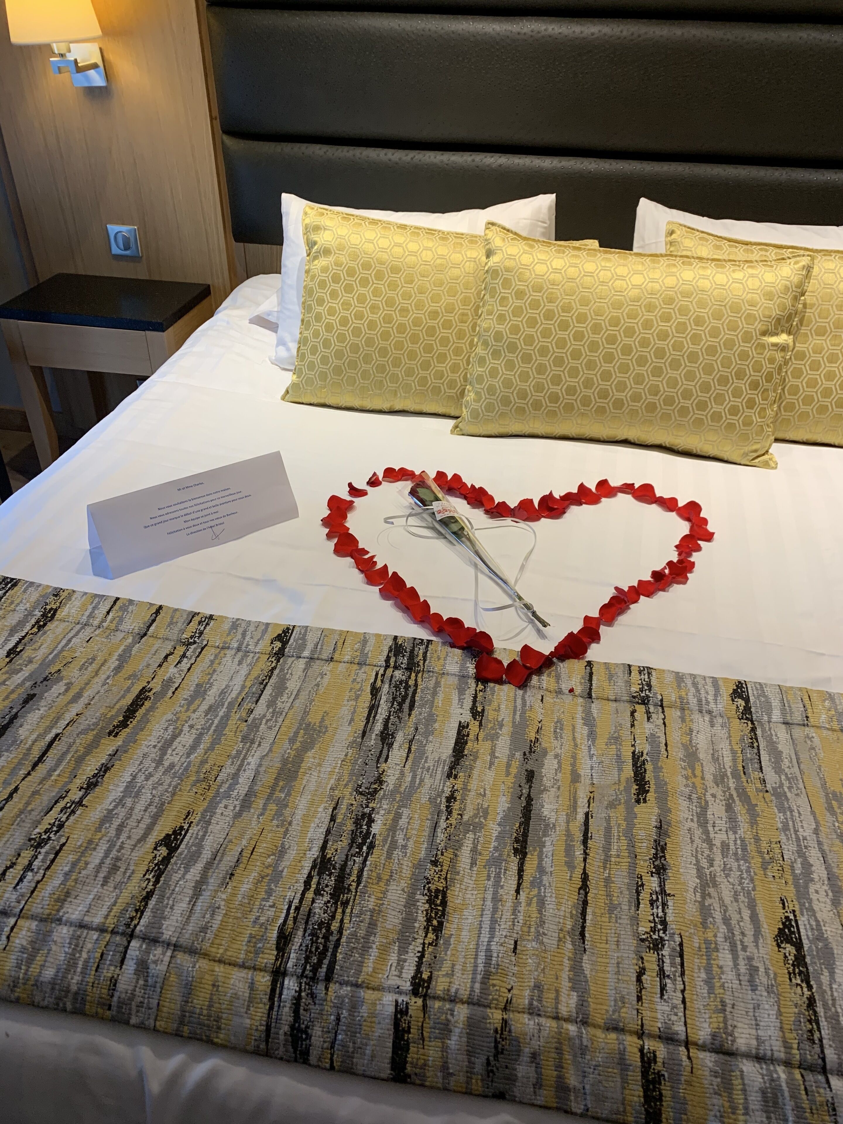 A neatly made bed with a heart-shaped arrangement of red rose petals and a bouquet of flowers, accompanied by a card.