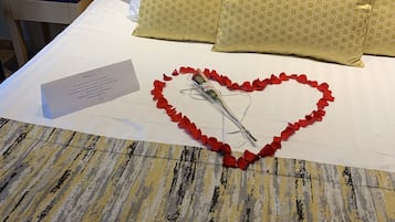 A neatly made bed with a heart-shaped arrangement of red rose petals and a bouquet of flowers, accompanied by a card.